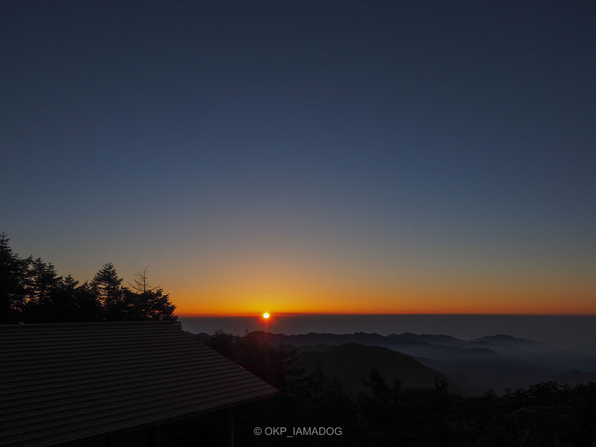 雲取山の山頂から見た朝日と奥多摩の山並みの写真。濃紺の空の下、オレンジ色に染まった雲の縁に朝日がかかっている。