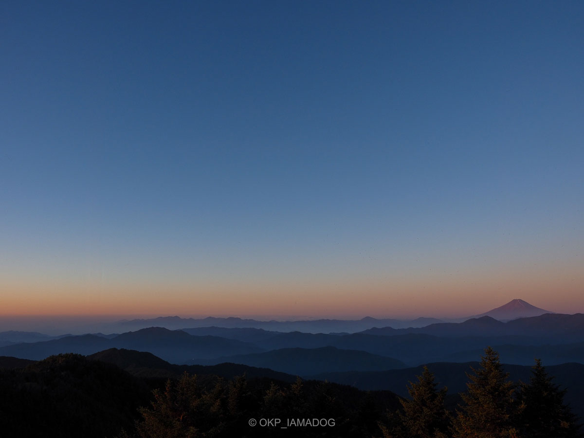 雲取山の山頂から見た奥多摩の山並みと夜明けの富士山の写真。まだ暗い空の下、朝日を受けた富士山が赤く浮かび上がっている。