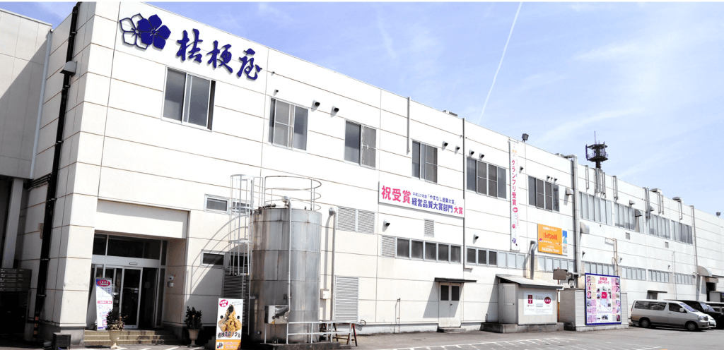 青空の下に立つ、白い壁の大きな工場。建物の上には「桂新堂」と書かれている。
