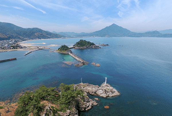 険しい岩山の先に灯台が見える、海沿いの町並みの空撮。