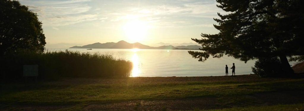 夕暮れの湖畔で、2人の人物が水面に映る夕日を眺めている。