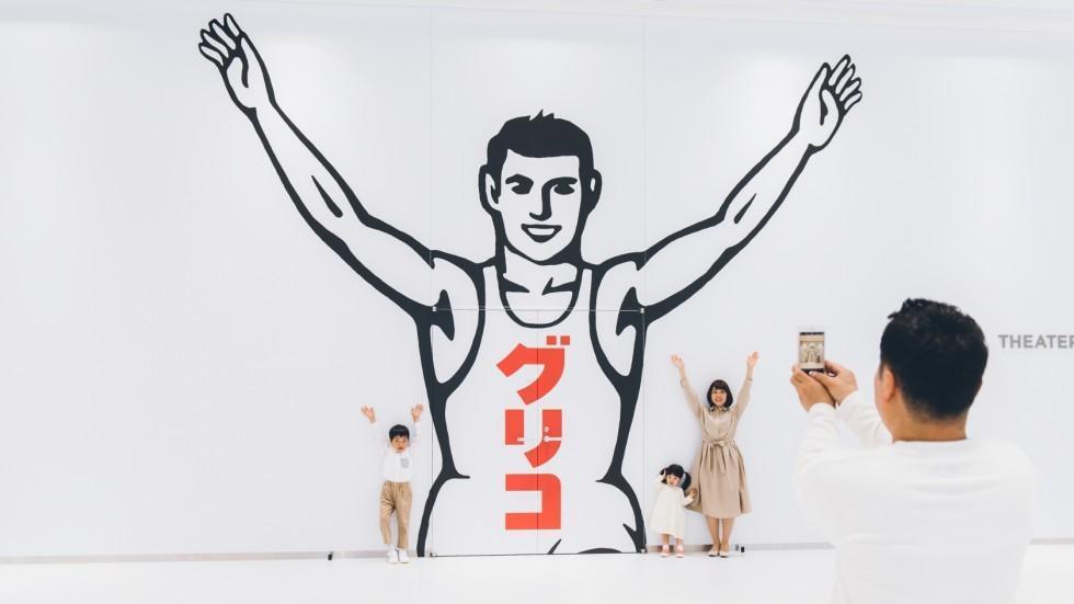 大きな「グリコ」のランナーのイラストの前で、家族が記念撮影をしている。