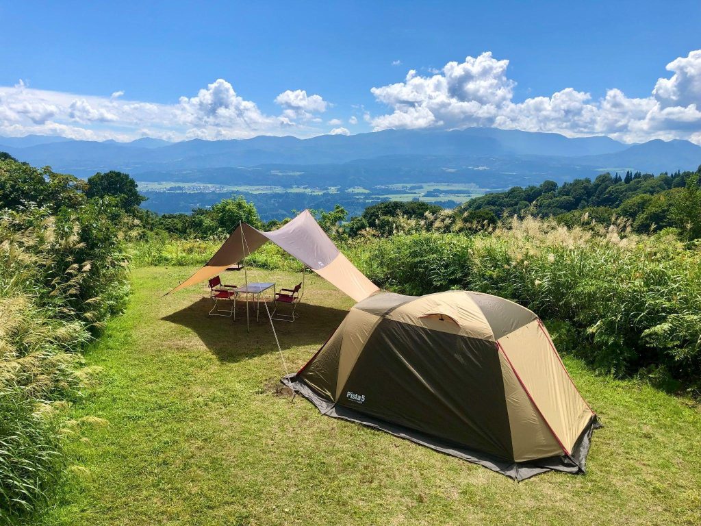 見晴らしの良い丘の上のキャンプ場。テントとタープが設営され、遠くに山々と街並みが見える。