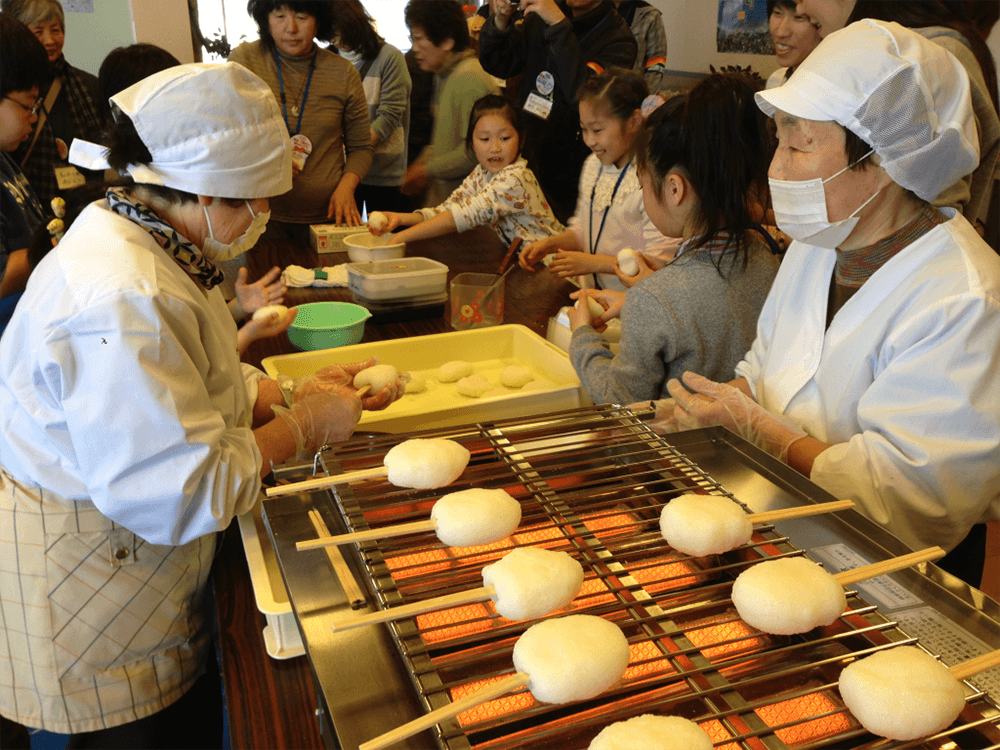 大人や子供たちが、串に刺したお餅を網の上で焼いている。