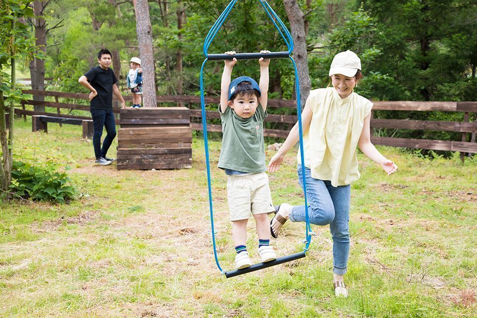 広場で遊ぶ家族。母親が子供をブランコに乗せ、父親と別の子供が見守っている。