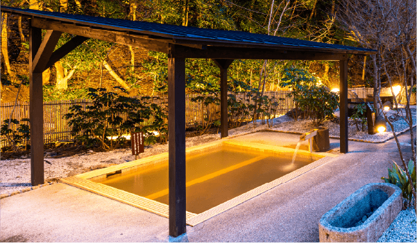 夜にライトアップされた、屋根付きの露天風呂。湯船の隣には、お湯を注ぐ木製の樋がある。