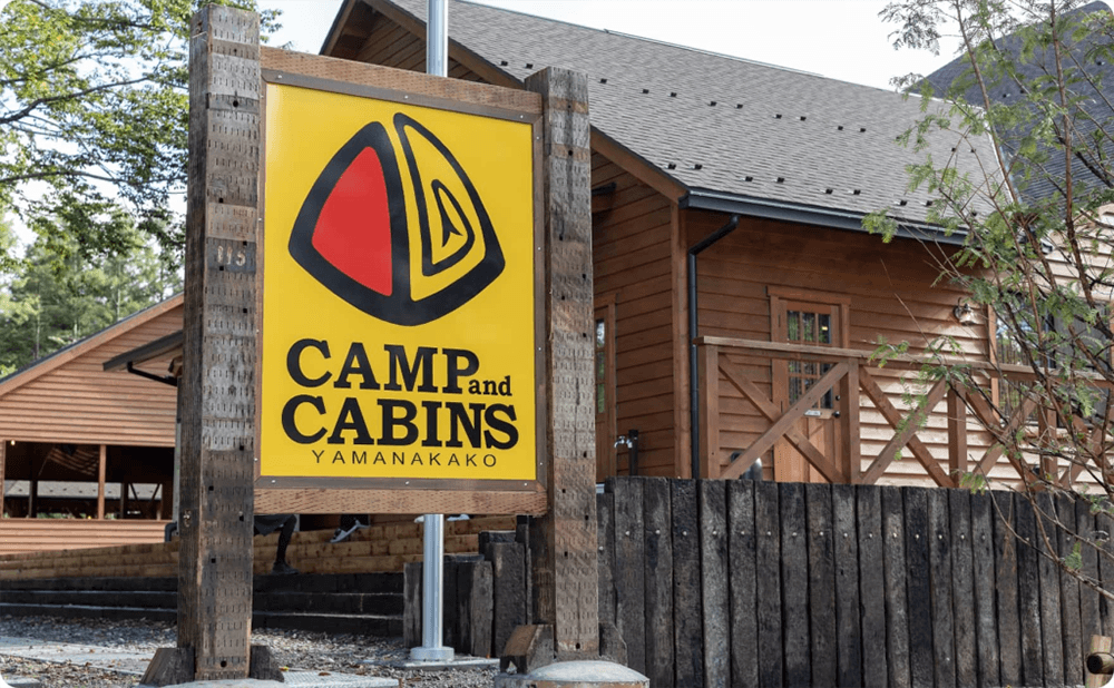 「CAMP and CABINS YAMANAKAKO」と書かれた黄色い看板と木造の建物。
