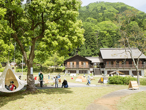 芝生の広場で遊ぶ人々。中央には木造の建物が建ち、木に吊るされたハンモック型の椅子に座る子供が見える。
