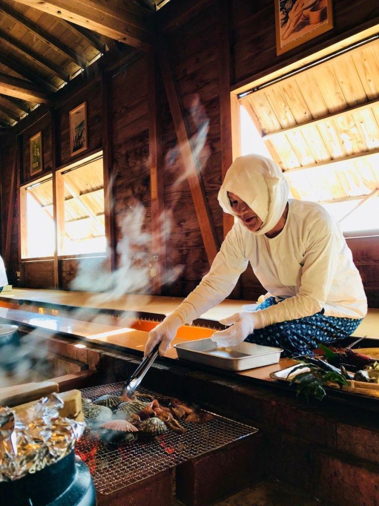 日本の伝統的な囲炉裏のそばで、ホタテなどの魚介類を網で焼いている人物。