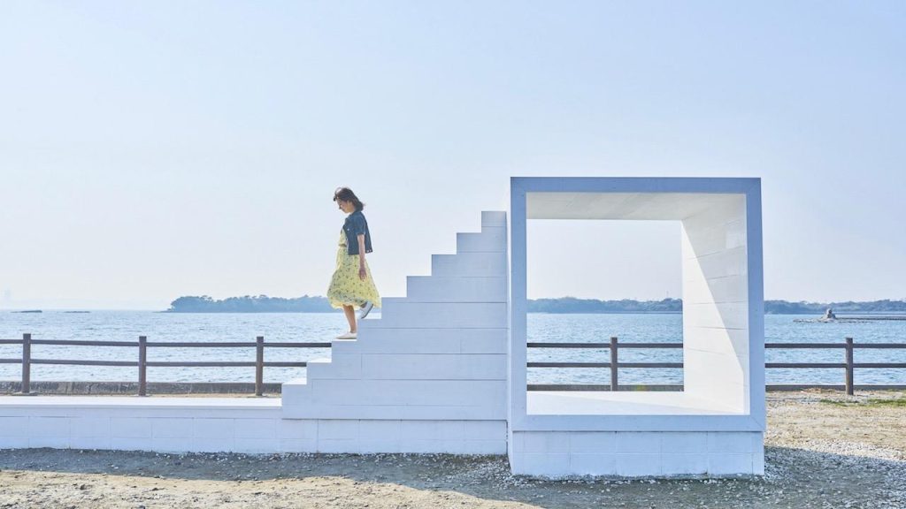 海を背景に、白いモダンな階段とフレームのオブジェ。階段を降りる女性が一人いる。