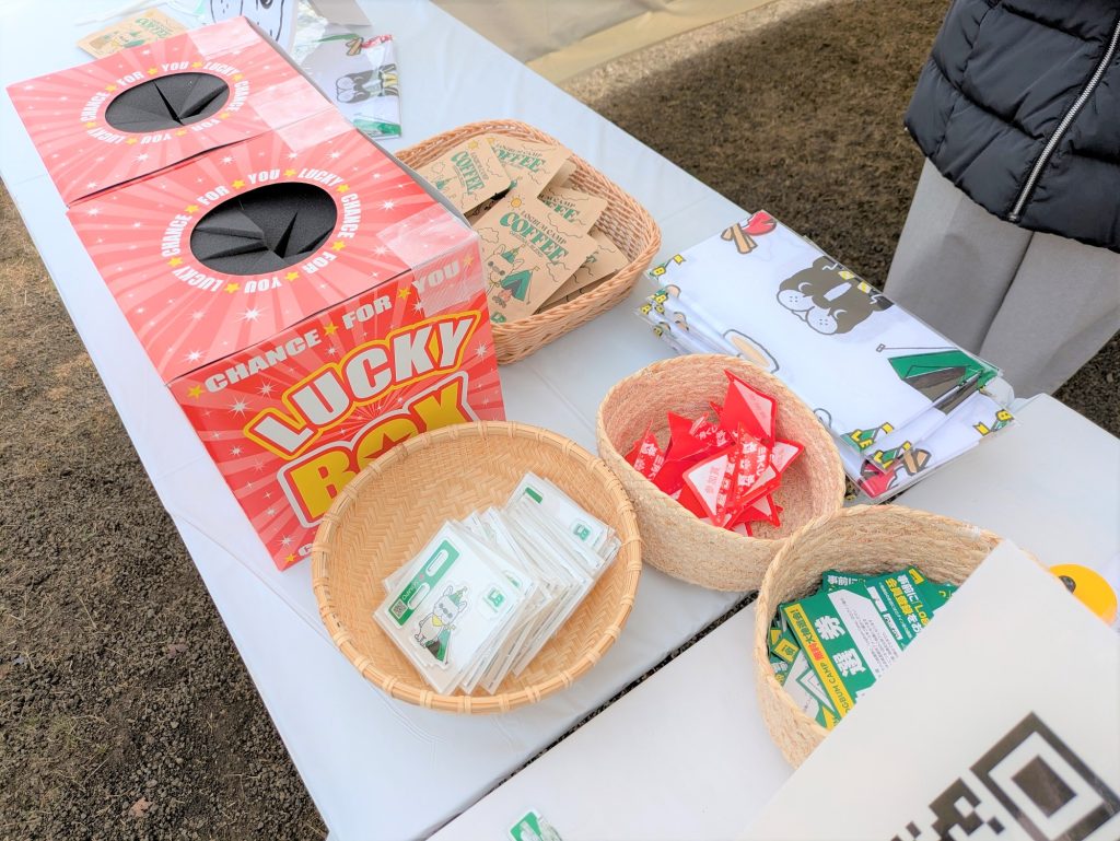ブースのテーブル。景品と思われる「LUCKY BOX」と書かれた赤い箱、コーヒーバッグ、くじ券、手ぬぐいが置かれている。