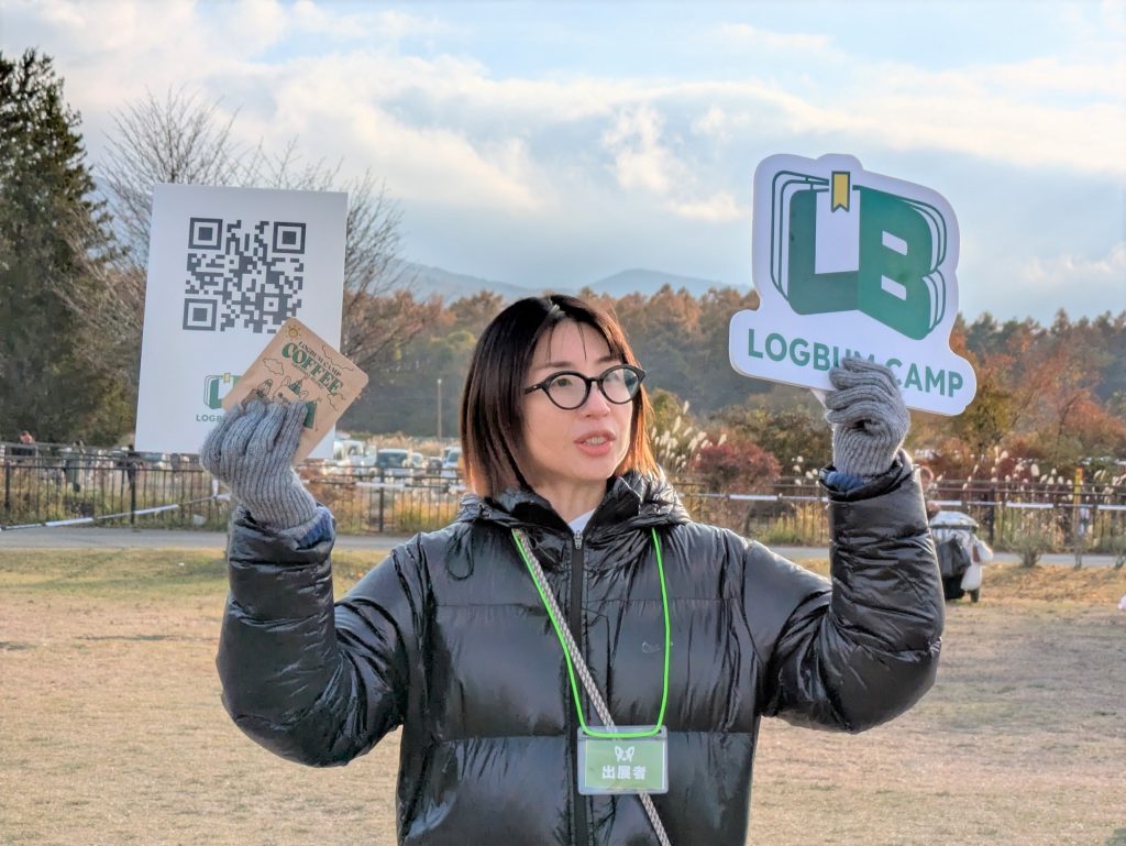 キャンプ場で、黒いダウンジャケットを着た女性が「LOGBUM CAMP」のロゴが入ったパネルとQRコードを掲げている。
